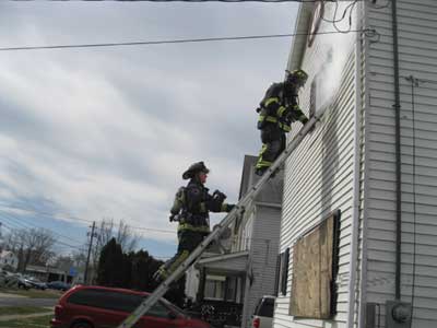 (2) The VES firefighter at the top of the ladder prepares to ventilate the window. The oriented firefighter below him is carrying the thermal imaging camera (TIC).