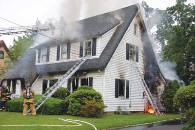 (2) The fire appears to be knocked down on the first floor, and the roof is being vented. Because of the peak of the roof, the roof firefighter is operating off of an aerial ladder.