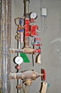 (2) A combination standpipe/sprinkler system. A pressure-reducing sprinkler floor control valve is tapped into the standpipe riser above a pressure-reducing 2½-inch hose outlet. Note the supervisory switch on the floor valve's hand wheel, the water flow switch, and the system side pressure gauge. The pressure-reducing floor valve keeps pressure in the sprinkler piping and heads from exceeding 165 psi. To further safeguard the system, a relief valve, located opposite the system pressure gauge, is set to open at 175 psi and dump water into drain piping, seen to the right of the riser.