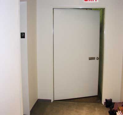 This stair door is unable to self-close because of severe winter stack on Floor 13 of a 100-story building. These open doors well below the NPP can pull fire/smoke toward occupied exit stairs, endangering evacuees on the floors above. Crews must check for these open doors to keep the stairway secure; stair pressurization is critical.