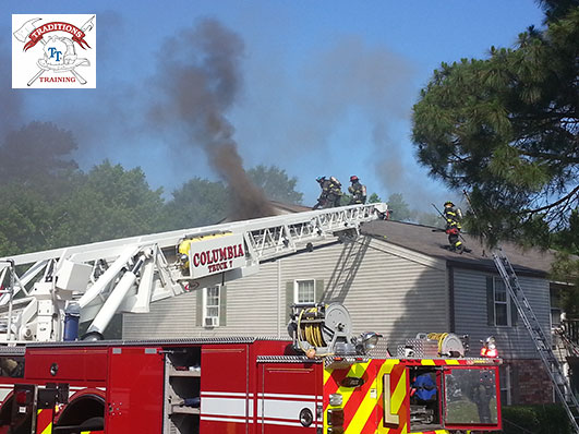 Aerial positioed at the scene of a fire, with firefighters on the roof of a home during a fire call. Smoke is venting from the upper portion of the house.