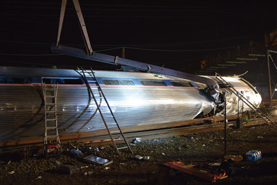 Car 2 lies on its side. A catenary pole penetrated its stainless-steel body. Portable battery-powered LED lighting illuminates the scene. Responders used numerous portable ladders to access the windows of each of the four overturned rail cars. Portable generators and hydraulic extrication equipment are staged outside the car.