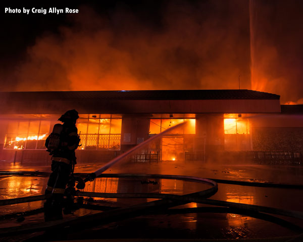 Flames shoot from the structure as a firefighter maintains an exterior hosestream on the fire.