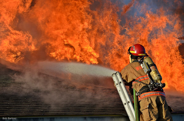 Flames erupt from the roof oa North Carolina home as a firefighter in full gear uses a hoseline to apply water while at the top of a ladder.