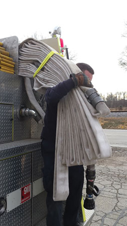 (6) A firefighter deploys the 100-foot flat load section of the DL Blitz Load. The strap is used to hold the load together on the firefighter’s shoulder; it eventually falls off when the load starts to deploy. Although the firefighter is carrying the 100-foot section away from the engine, the second 150-foot section is being deployed, and no mess occurs behind the engine. The load is lightweight, maneuverable, and quickly deployed. 