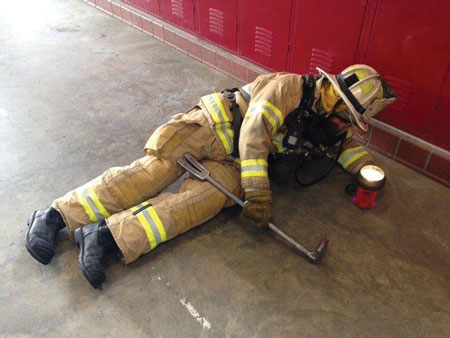 (2) The firefighter has positioned his body perpendicular to the wall to better allow search crews to find him. He shines his light to the ceiling in flashing mode and uses his leg to support the tool while striking it on the ground. Using your leg as a fulcrum allows you to save upper body strength by not having to support the full weight of the tool. Remember, if you know the location of the doorway, point your flashlight in that direction to alert incoming crews.