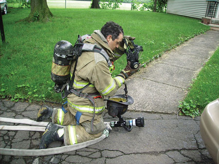 <b>(21) </b>The nozzle firefighter secures the nozzle and the coupling as he dons his self-contained breathing apparatus face piece.<i> (Photo by Michael Clark.)</i>