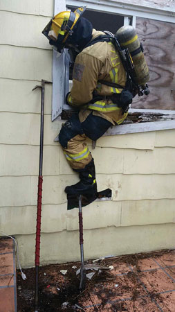 (6) The search position FF is making entry at this first-floor window. For training purposes, only half of the window was used to prevent further damage to this structure. Note that the roof hook is placed near the window to be used as a reference point from the windowsill and the halligan bar is used as a step to assist in making the entry.