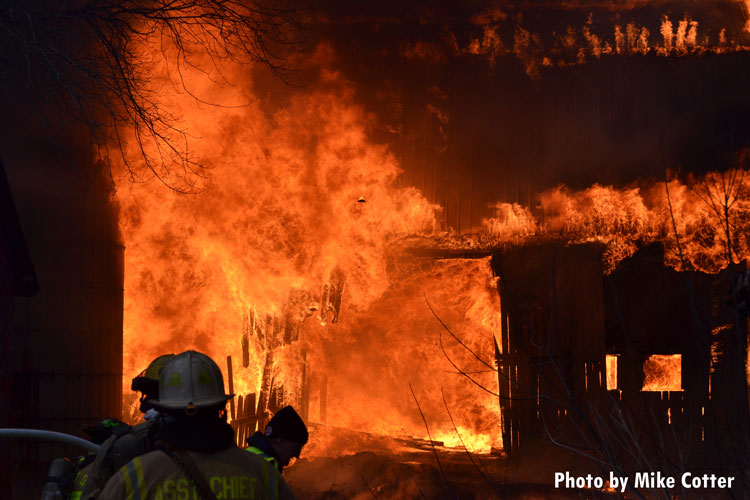 Flames rage in the background as firefighters ready to apply water at a live burn in a barn.