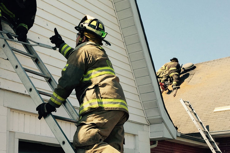 A firefighter reaches up a ladder while others perform roof work.