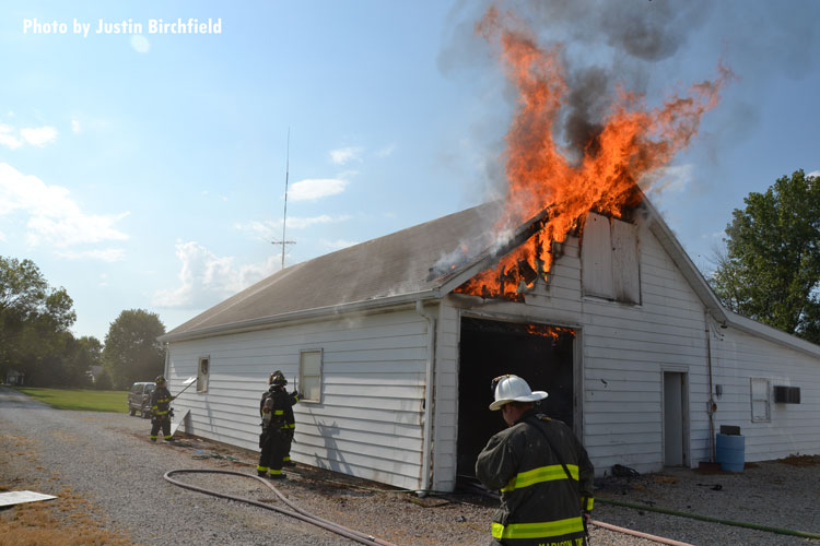 Fire vents from the roof of a barn as firefighters prepare to attack