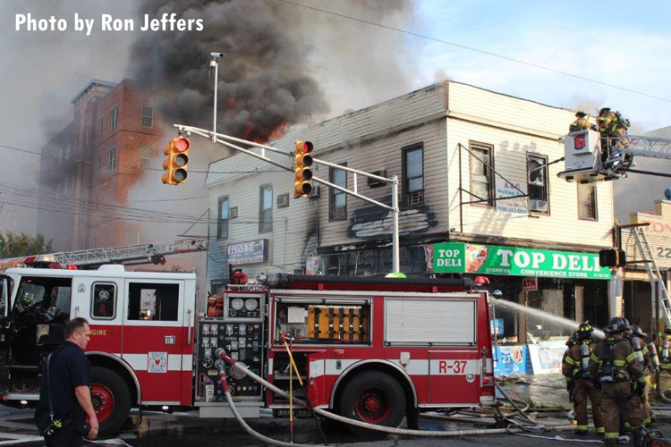 Fire vents through the top of a building in Jersey City as firefighters operate in a tower ladder bucket and hoselines are stretched from a fire engine