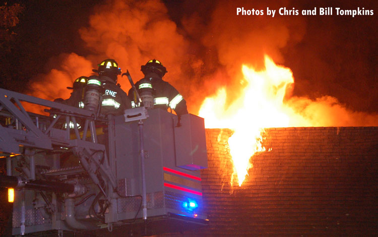 Firefighters in a bucket confront flames shooting from the roof of a home