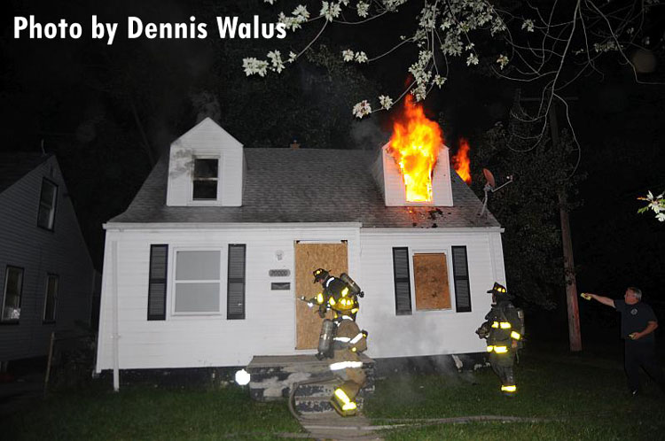 Detroit firefighters operating at a house fire with flames venting from upper window