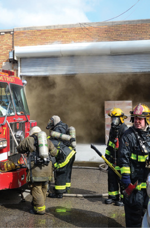 (2) Additional personnel arrive to assist interior crews. When crews are operating in enclosed structures, ensure that staffing is sufficient or request additional personnel. A large overhead rolling door provides an excellent opening for access to the fire and ventilation. At the upper left of the doorway, powerful torsion springs that counterbalance the door’s heavy weight assist the chain hoist mechanism. Heat attacking the torsion springs at the top of the doorway can cause them to lose their tension and the door may close unexpectedly, trapping firefighters in the building. Before making entry, it is very important to brace the door in the open position with a pike pole or other means. 