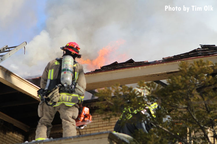 A firefighter with a saw stares up at flames venting through a roof during a house fire