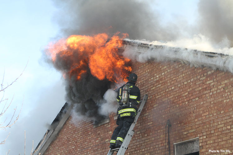 Firefighter on a ground ladder as flames vent through an upper window