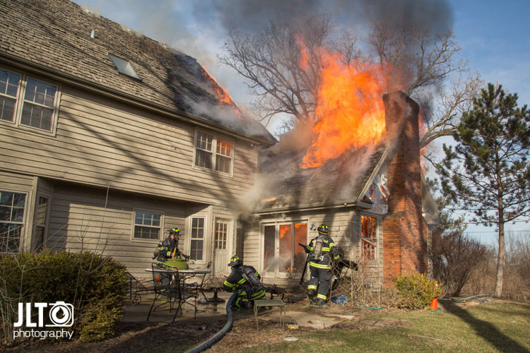 Flames erupt from the roof of a home as firefighters operate