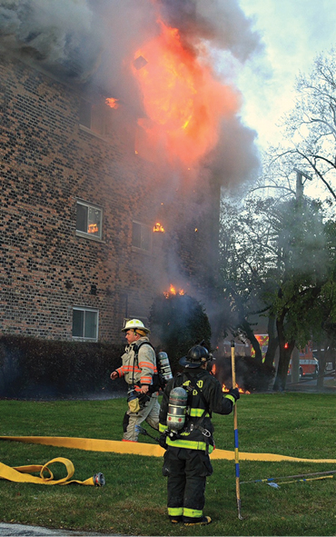 (2) Debris is on fire and falling from the roof overhang. Large-diameter hose is being moved to supply an elevated stream.