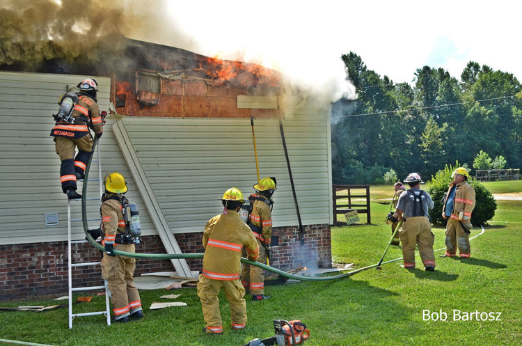 Firefighters operate a hoseline from a ground ladder as flames emerge from the eaves of a home