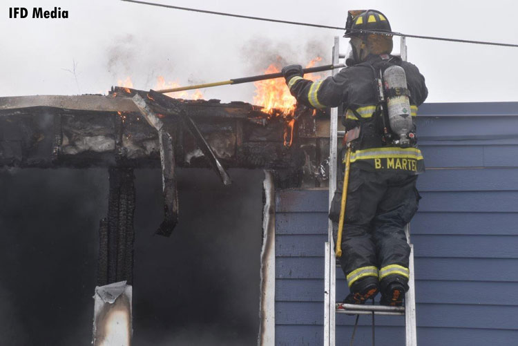 Firefighter on a ladder with a pike pole with flames showing from roof