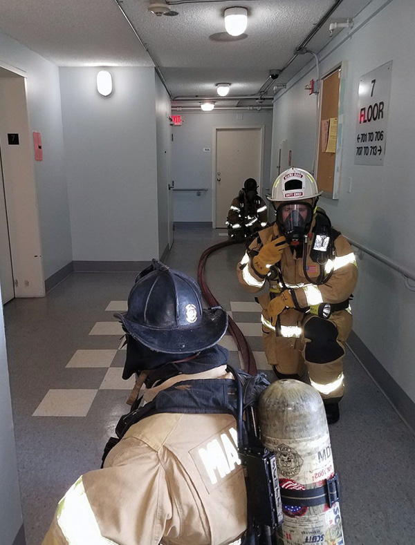 Firefighters maneuver hose around corners in an elevator lobby. They are being directed by a battalion chief who, as fire floor division supervisor, is not physically involved in the task.