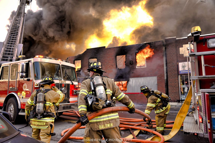 Firefighters manage hoselines in the street in front of a burning building