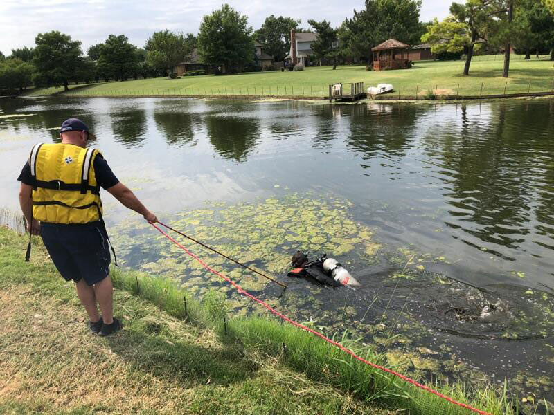 A diver navigates algae atop the pond while member on shore bearing a rope looks on