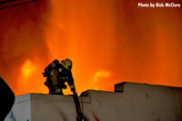 A firefighter on the roof of a structure with flames raging behind him.