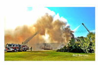 Fire apparatus and firefighters are enveloped in smoke from the burning structure.