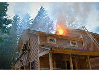 A firefighter on a ladder as fire vents from an upper floor.