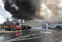 A tower ladder and firefighters operating a handline on the exterior of the building as flames and smoke vent from the roof