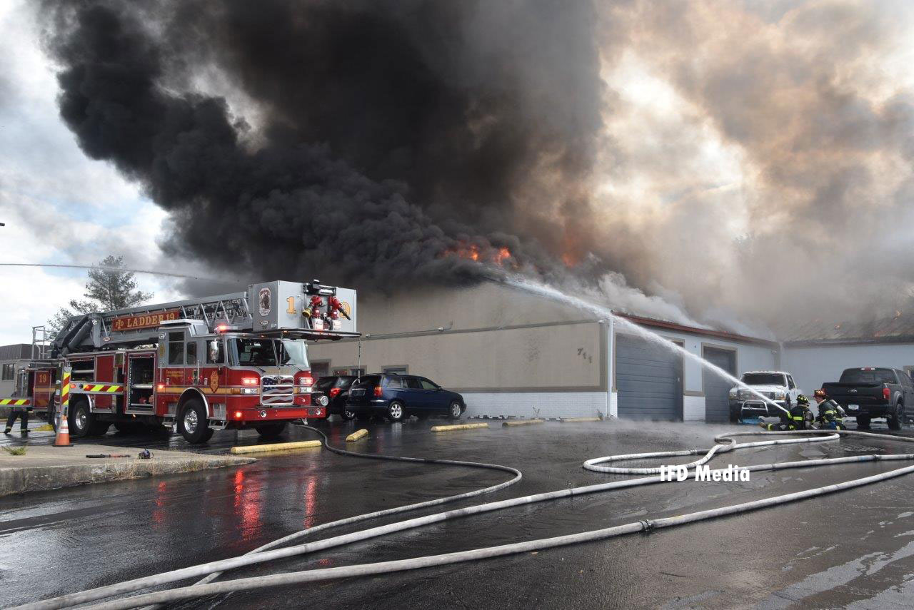 A tower ladder and firefighters operating a handline on the exterior of the building as flames and smoke vent from the roof