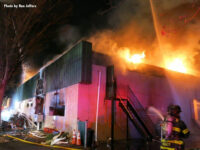 Fire burns through the building's roof as firefighters apply water.