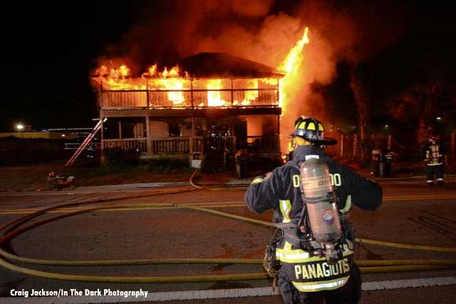 A firefighter wearing SCBA with hoselines and a home in fire behind him