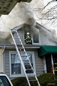 A firefighter on the exterior of a roof while smoke shoots from an upper floor