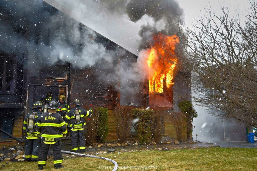 Fire pours from a window as firefighters are poised outside the structure with hoselines