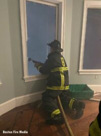 A firefighter on the interior of a dwelling directs a hose stream through a window