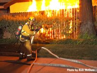 Firefighter with a hoseline near a fence as fire rages in a building in the background