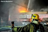 An LAFD firefighter with a TIC as flames shoot from a building in the background