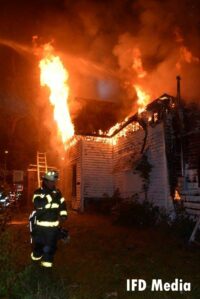 Firefighter at the scene of an arson fire as flames shoot from underneath a roof