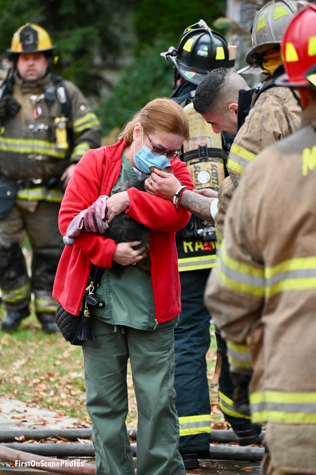 A resident cuddles a dog that firefighters rescued from a fire as firefighters look on