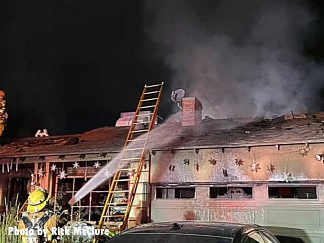 A ladder raised with a firefighter training a hose stream on the top of a home