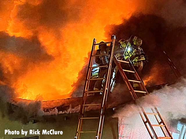 Two ladders nearly meeting atop a roof at a house fire with firefighters on the roof