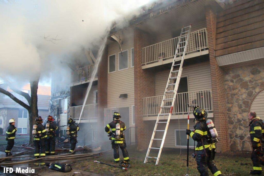 Firefighter puts a hose stream on the roof of an apartment fire with ladder extended up the balcony