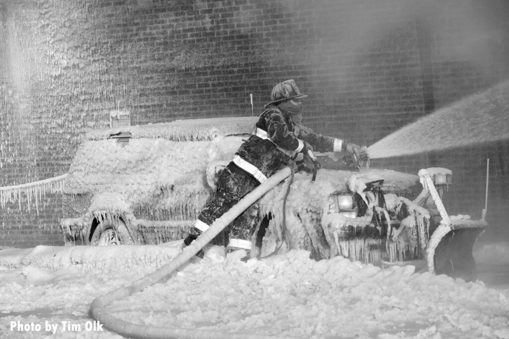 Firefighter leaning over a car covered with icicles with a portable master stream