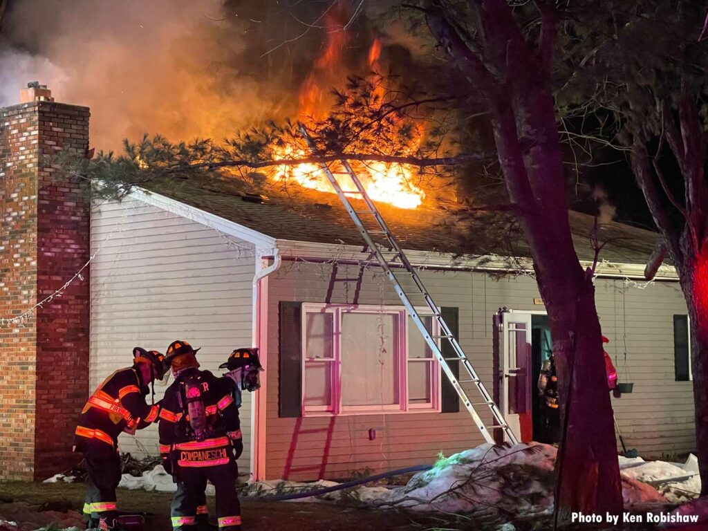 Flames shoot from the roof as firefighters work, with a ground ladder slung against a building