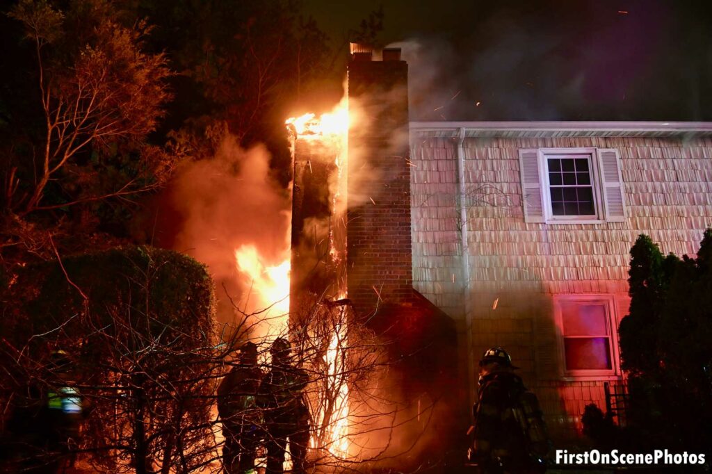 Firefighters at the scene as flames leap up the exterior walls during a Long Island house fire