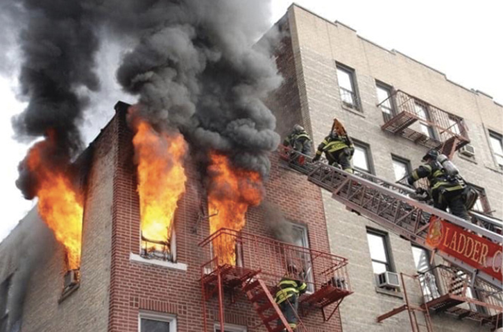 The outside vent firefighter patiently waits on the fire escape to vent the top-floor window as roof firefighters ascend the aerial directly above.