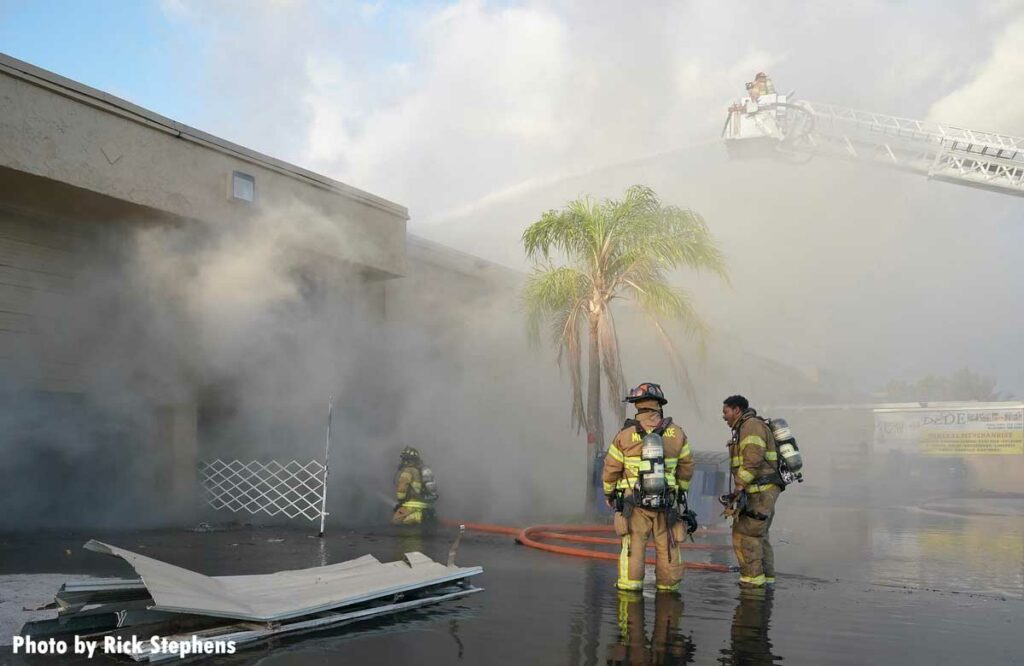 Firefighters in tower ladder put water on burning warehouse with other firefighter in foreground